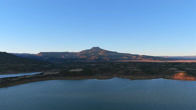 A wide drone view shows a flat-top mountain overlooking a large blue lake at sunset. This high-desert scene in Abiquiu, New Mexico, features golden light and rugged western beauty.