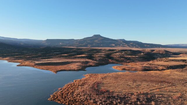 Wide aerial view shows blue water of Abiquiu Lake reservoir meeting arid, golden brush shorelines. The volcanic basalt cap of Cerro Pedernal mountain dominates the high desert plateau horizon.