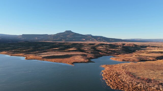 The blue water of Abiquiu Lake reservoir sits below the volcanic basalt peak of Cerro Pedernal. The high desert plateau landscape features sunlit soil and arid shorelines at the base of the mountain.
