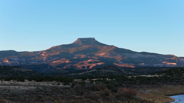 Low-angle sunlight illuminates the jagged peak of an ancient flat-top mesa in New Mexico. This detailed view captures the solitary mountain rising above a blue lake in the vast high desert