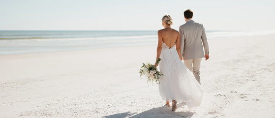Newlywed couple walks along pristine shoreline in elegant wedding attire holding bridal bouquet.