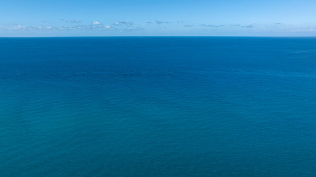 A serene wide shot of the deep blue Sea, with a clear horizon line under a soft blue sky and distant faint clouds. Aerial view of the vast ocean.