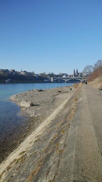 Vertical static shot along the concrete Rhine embankment and promenade in Basel, Switzerland, featuring a pebbled beach in the foreground and the historic Basel Minster with Wettsteinbr&uuml;cke in the dis