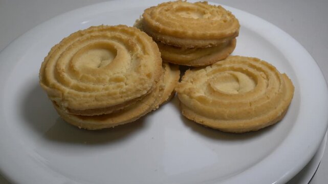 A small stack of round butter cookies with a spiral pattern rotates slowly on a white plate, showcasing their crumbly texture and golden-baked edges in a studio setting.