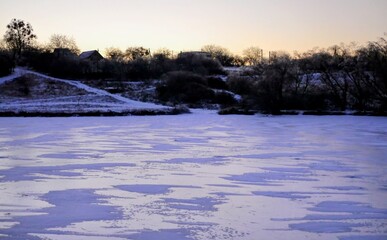 Obraz premium Winter landscape with a frozen lake, snow covered lake bank, and silhouettes of rural houses on the horizon. 