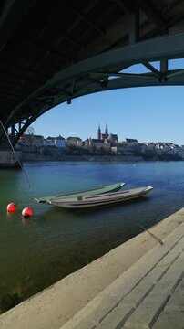 Vertical static shot from under the steel arch of the Wettsteinbr&uuml;cke, featuring traditional wooden Weidling boats on the Rhine river with the historic Basel Minster and old town skyline in the backgr