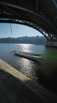 Vertical static shot from the riverbank looking under the dark steel arch of the Wettsteinbr&uuml;cke bridge, featuring traditional long wooden boats (Weidling) moored on the sparkling Rhine river in Basel