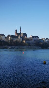 Vertical static shot of the iconic Basel Minster (Basler M&uuml;nster) with its twin towers on the hill overlooking the blue Rhine river under a clear sky in Basel, Switzerland.