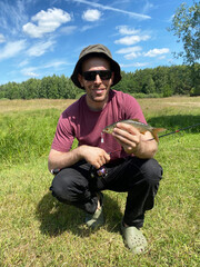 Man angler wearing sunglasses and a hat holds a freshly caught fish while kneeling on grass in a sunny outdoor setting with trees in the background