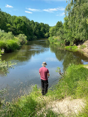 Man fishing by a calm river surrounded by lush green trees and grass under a clear blue sky, wearing a hat and standing on the riverbank