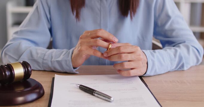 Lawyers client thinking about divorce in front of marriage annulment paper document. Woman sitting at wooden desk with judge gavel over court paperwork, taking off and putting on wedding ring