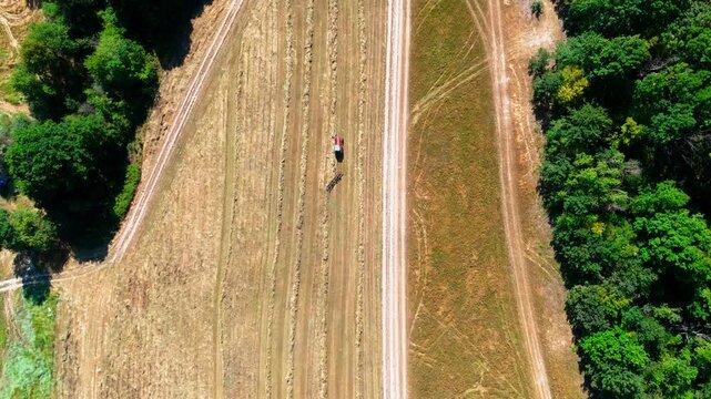High-altitude overhead drone footage of a red tractor raking straw for hay bales in a cultivated field, bordered by dense green forest on both sides, showcasing agricultural work and rural patterns.