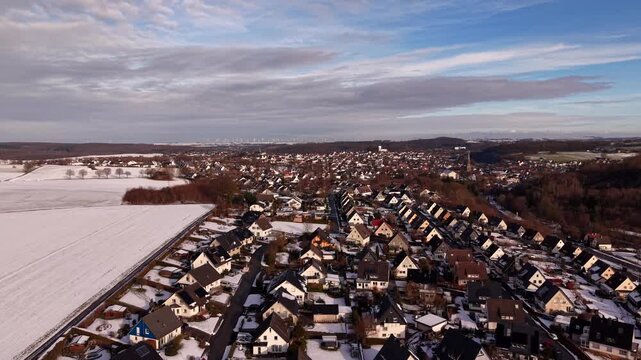 Aerial view of snow-covered residential neighborhood in Warstein, Germany, under winter sunlight, with clustered houses, icy rooftops, surrounding fields and calm seasonal atmosphere. Wide shot.