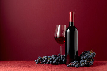 Bottle and glass of red wine with blue grapes on a red background.