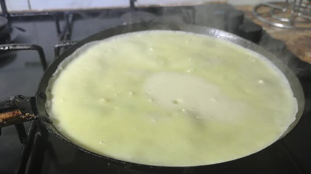 A person pours batter onto a hot pan to make dosa in a home kitchen. The process shows how to cook this dish using basic tools and a stove.