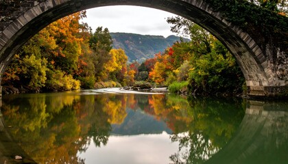 River reflects colorful autumn trees under a stone bridge arch, with hills in background, under a cloudy sky