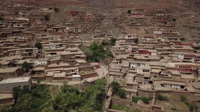 Aerial drone shot of an indigenous Berber village in the High Atlas. Features include traditional earth architecture, vibrant green valley, and weathered red geological mountain formations.