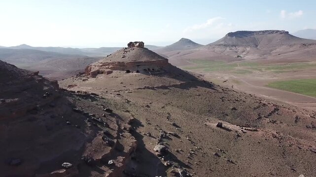 Aerial drone shot of a unique mountain summit in the High Atlas. Features include natural geological caves, ancient human-adapted rock shelters, and rugged prehistoric textures in Morocco.