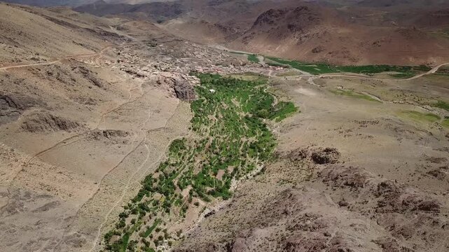 Aerial drone shot capturing the contrast between fertile green alluvial patchwork and arid mountain strata. Highlights include rugged rock formations and a dry riverbed in the High Atlas.