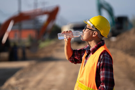 Tired engineer sitting on ground at construction site, feeling hot and thirsty.
