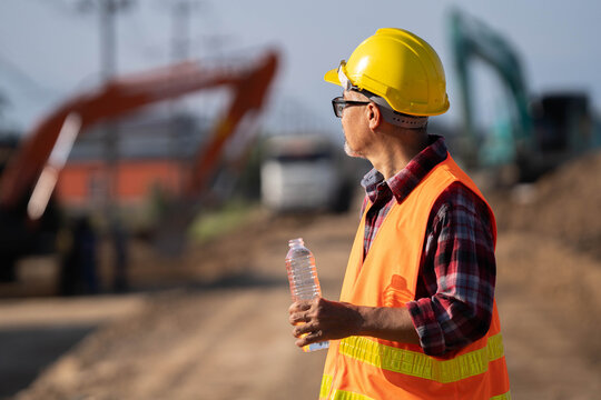 Tired engineer sitting on ground at construction site, feeling hot and thirsty.