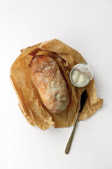 Ciabatta bread, cream cheese, and a butter knife are placed on a brown paper wrapper. White background.