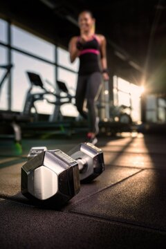 Metal dumbbell lying on floor of gym, female athlete training in background in sunlight from large window. Concept of fitness, strength training, building muscle and achieving goals.