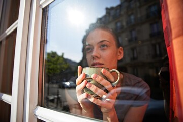 Serene young woman holding mug of coffee, looking out the window at historical big cityscape, drinking, daydreaming, enjoying peaceful moment.
