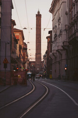 Colorful buildings line the narrow streets of Bologna as the sun sets, casting a warm glow on the towering landmark. People walk leisurely, enjoying the vibrant atmosphere of this Tuscan gem