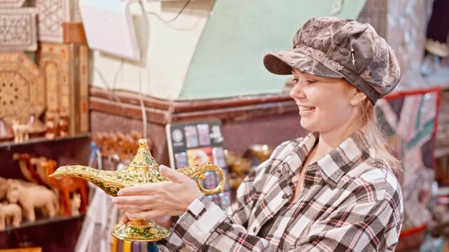 Smiling woman holding a decorative Arabian magic lamp at a souvenir market. Travel shopping concept, Middle Eastern culture, tourism, tradition, and curiosity.