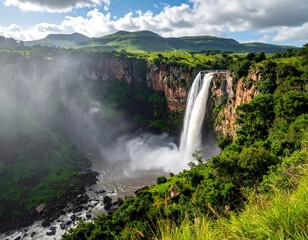 Waterfall plunges from cliff face in verdant landscape with mountain backdrop under a bright blue, partly cloudy sky