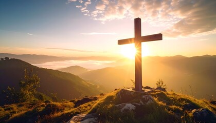 Wooden cross atop a grassy hill against a mountain sunset, bathed in warm light and with cloudy skies
