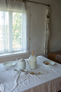 Traditional breakfast table setting with white dishes, teapot, and pitcher by a bright window in a rustic room