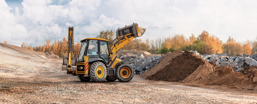 A loader works on a construction site, lifting dirt and rocks near a pile of debris. Bright clouds fill the sky. Autumn trees are visible in the background