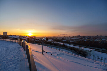 Fototapeta premium Panoramic winter cityscape viewed from a hill. A snow-covered highway interchange with sweeping curves leads toward a distant city of mid-rise buildings. To the right, a wide frozen river stretches al