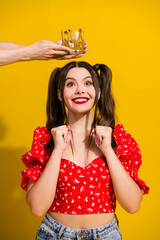 Young glamorous girl smiles as crown is placed on her head against a yellow backdrop in a playful...