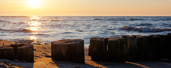 Naklejka premium Wooden breakwater stretches into blue sea under soft evening light. Gentle waves touch sandy shore while sun reflects warmly on weathered timber posts and shimmering water