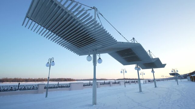 A wide-angle, low-angle shot of a snow-covered river promenade in winter. A striking modern, slatted metal canopy is suspended by cables over the walkway. The path is lined with street lamps and borde