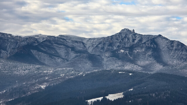 Panoramic view of the Ceahlău Massif in Romania under a cloudy sky. Toaca Peak rises above snow-dusted pine forests and rugged Carpathian mountain ridges in a serene winter landscape.
