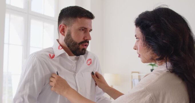 Suspicion of adultery and infidelity betrayal of sad wife looking at evidence of deception on husbands shirt. Jealous upset woman pointing out red lipstick stains to man and questioning honesty