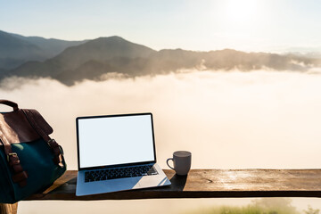 Fototapeta premium Empty screen laptop with cup of coffee and backpack with mountain view at sunrise in the morning
