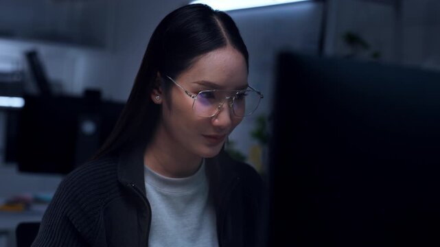 Happy young Asian woman female person wearing glasses eyeglasses looking computer screen monitor with smile working late night dark office feeling productive success joy then stressed tired headache
