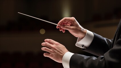 Male conductor in black tuxedo gestures with baton while leading an orchestra performance on stage with blurred audience in the background
