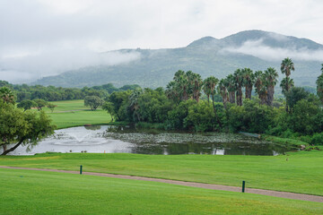 South Africa,Sun City,A serene golf course with a pond, lush greenery, and mist-covered mountains in the background. © SHUNJI ZHOU