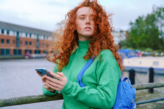 Young woman with curly red hair uses smartphone by the canal in a city on a cloudy day