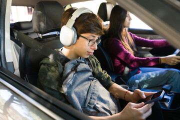 Teenager wearing headphones and glasses, using a smartphone in a car while being driven by a parent to school © AntonioDiaz