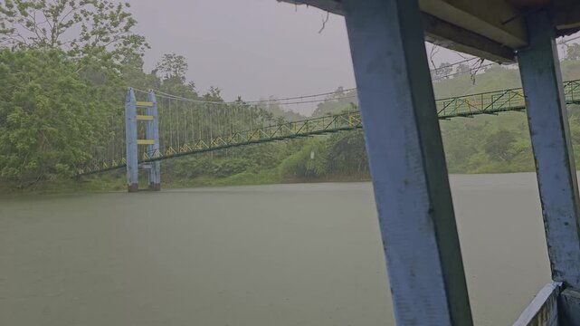 POV boat ride passing under metal suspension bridge on Dumboor Lake. View from inside wooden tourist boat with bamboo roof pillars during rain in Tripura.