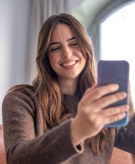 Fototapeta premium Portrait of smiling woman in video call to friends or relatives, using phone webcam, sitting on couch at home, happy young female blogger recording vlog, chatting online