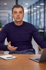 Fototapeta premium Man sitting at a desk in a modern office, speaking and gesturing during a conversation or presentation with a laptop and notebook in front of him