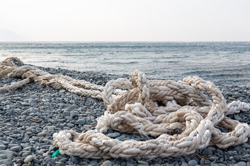 Fototapeta premium The ship's rope is in selective focus. Woven with thick bundles of threads, the ship's rope lies on a pebble beach. Türkiye, Samandağ, Hatay, Mediterranean coast.